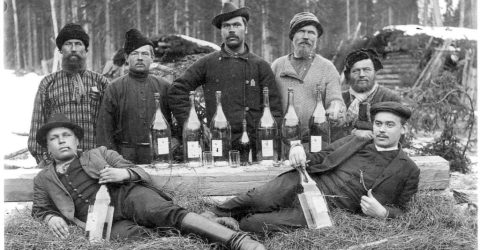 A group of Russian loggers pose around a fallen log and several bottles of vodka.
