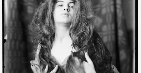1890 photograph of a young woman with her hair down, standning before a floral fabric backdrop.
