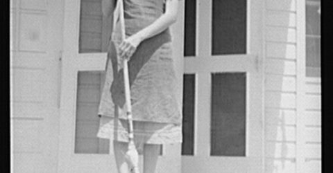 A youg woman sweeps the porch in front of a white clapboard house.